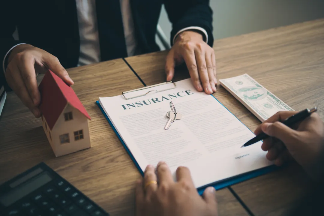 A person signs an insurance document with a pen, alongside a small house model and a dollar bill on a wooden table.